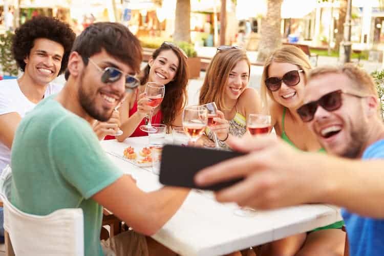 A group of six young adults sit around an outdoor table, smiling and holding drinks while taking a selfie together on a sunny day.